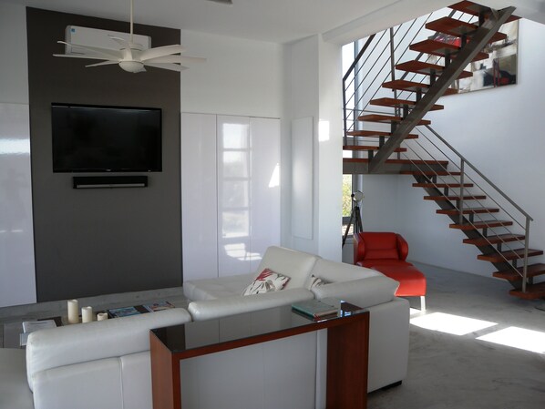 Una sala de estar moderna con un televisor de pantalla plana, un sofá blanco, una mesa de centro de vidrio y una escalera con escalones de madera.