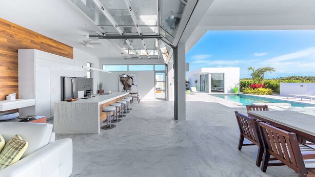 A modern kitchen with a central island, a dining area with wooden chairs, and a view of a swimming pool. - White Villas (Providenciales)