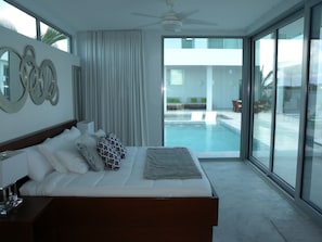 A modern hotel room with a large bed, a ceiling fan, a view of a swimming pool, and a balcony with outdoor seating.