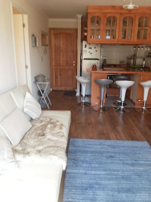 A living room with a sofa, a coffee table, and a rug. There is a kitchen area with wooden cabinets and a refrigerator. - Parque el Volcán (Pucón)