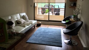 Sala de estar com um sofá branco, uma poltrona preta, um tapete azul e vista para carros estacionados através de uma janela grande.