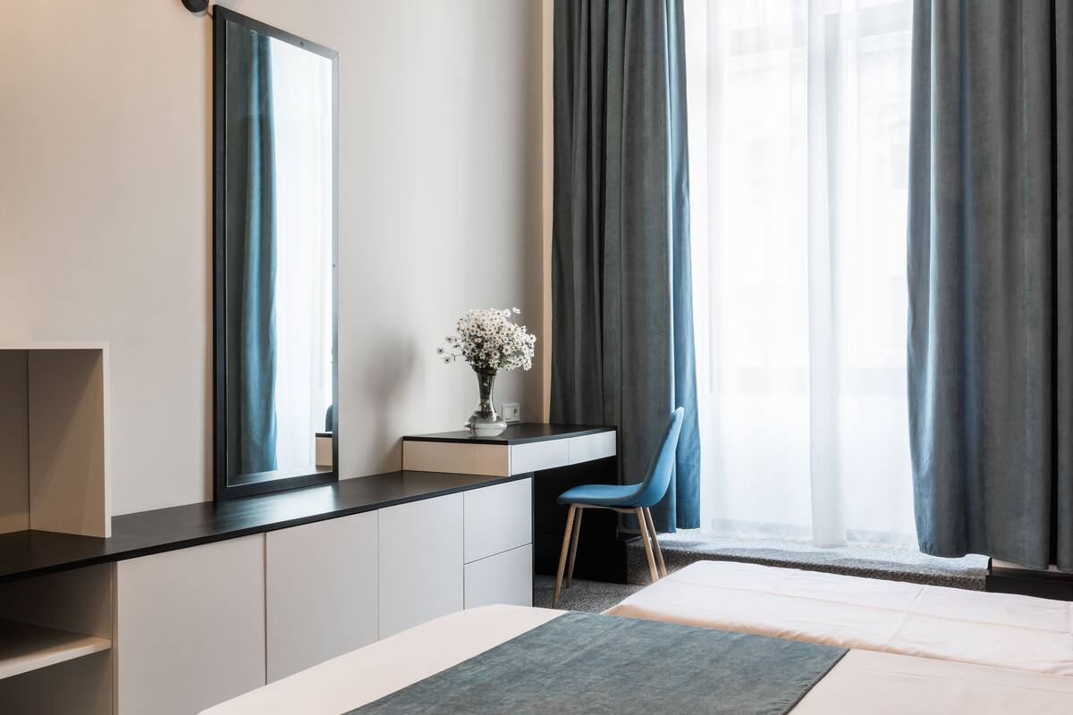 Une chambre d’hôtel moderne, équipée d’un lit, d’un bureau avec une chaise, d’un grand miroir et d’une fenêtre avec des rideaux.
