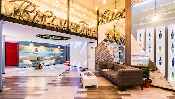 Eine moderne Lobby mit Fischgrätparkett, einem weißen Couchtisch, einem braunen Sofa und einer Wand mit gerahmten Weinflaschen.