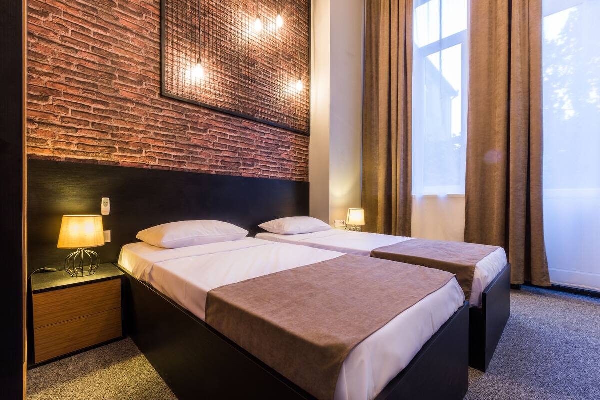 Une chambre d’hôtel avec deux lits, un mur en briques, une fenêtre avec des rideaux et une table de chevet avec une lampe.