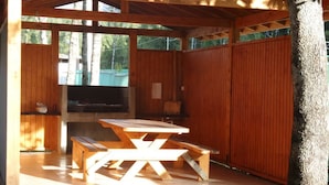 Un espacio al aire libre techado con una mesa de picnic y bancos, rodeado de paredes de madera y un árbol.