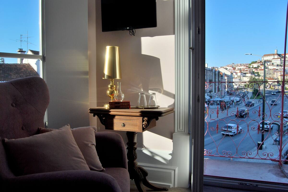 A room with a view of a busy street, a table with a lamp, and a sofa with cushions.