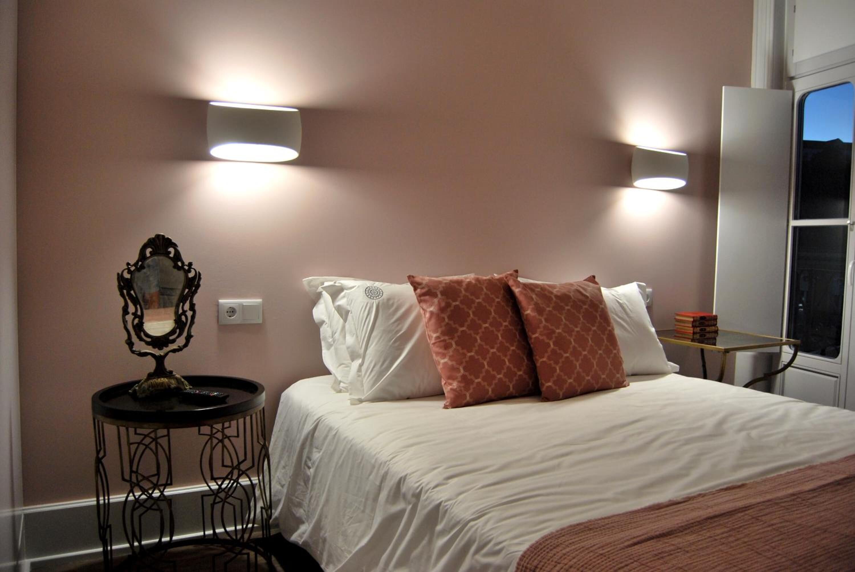 Un dormitorio con cama, mesita de noche con espejo y luces en la pared.