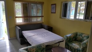 A compact room with a bed, a small table, and two chairs. There are windows with blinds, a potted plant, and a framed picture on the wall. - The Tranquil  C's (Nassau)