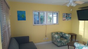 A room with yellow walls, a window with blinds, a ceiling fan, a sofa, a floral armchair, a small table, and a TV mounted on the wall. - The Tranquil  C's (Nassau)