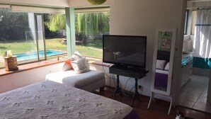 Un dormitorio con cama, televisor, una mesita y ventana con vistas a un patio trasero con piscina.
