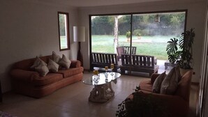 Salón con sofá marrón, una mesa de centro de cristal y vistas a un jardín a través de amplios ventanales.