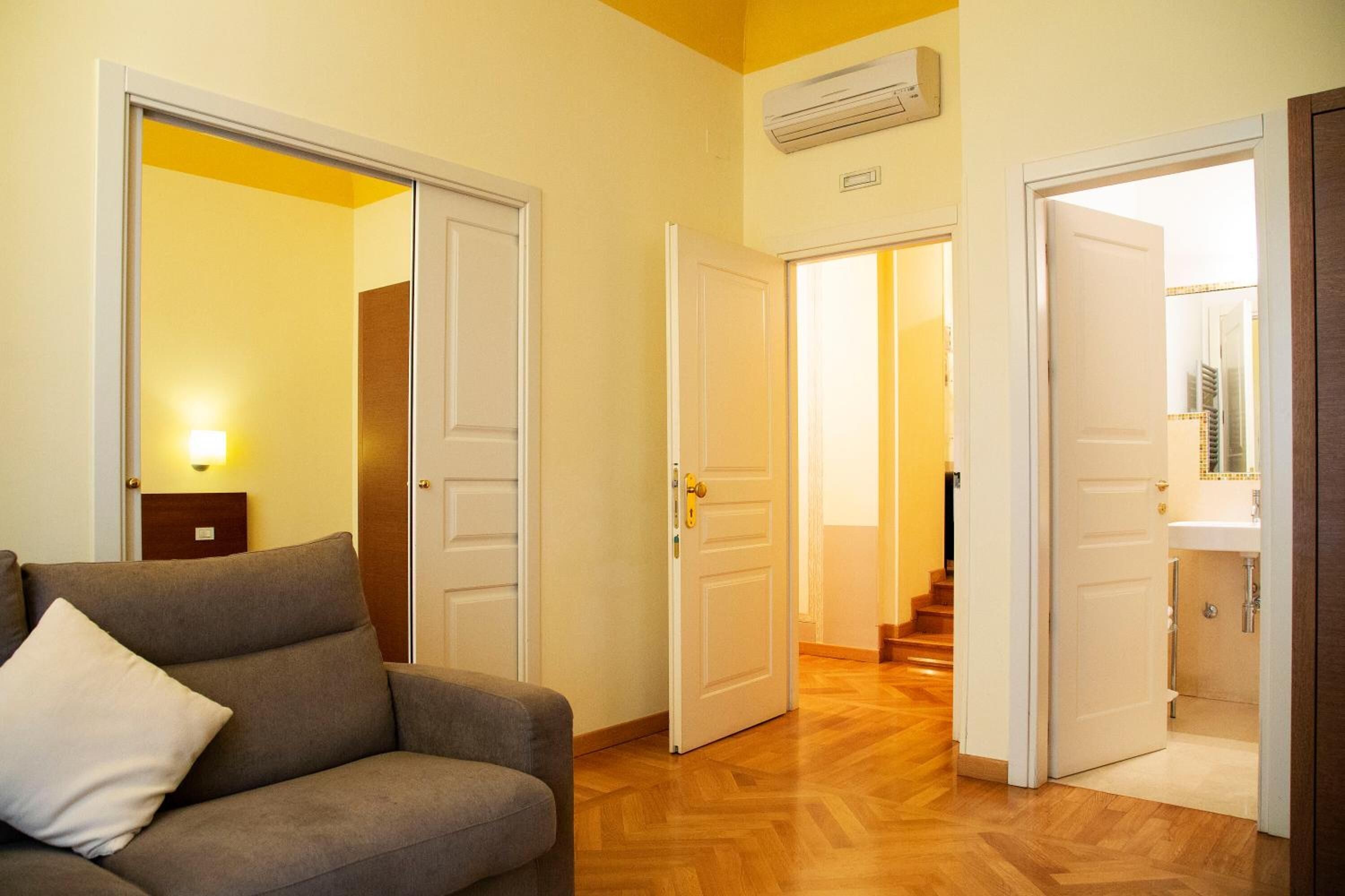 A hotel room with a sofa, a hallway with wooden flooring, and a bathroom visible through an open door.