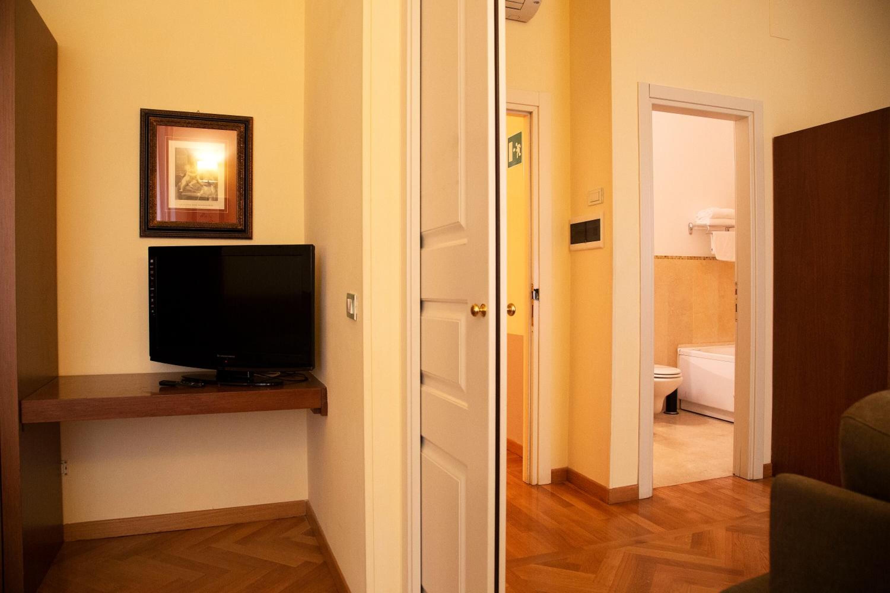 A hallway with a television on a shelf, a door leading to a bathroom, and a framed picture on the wall.