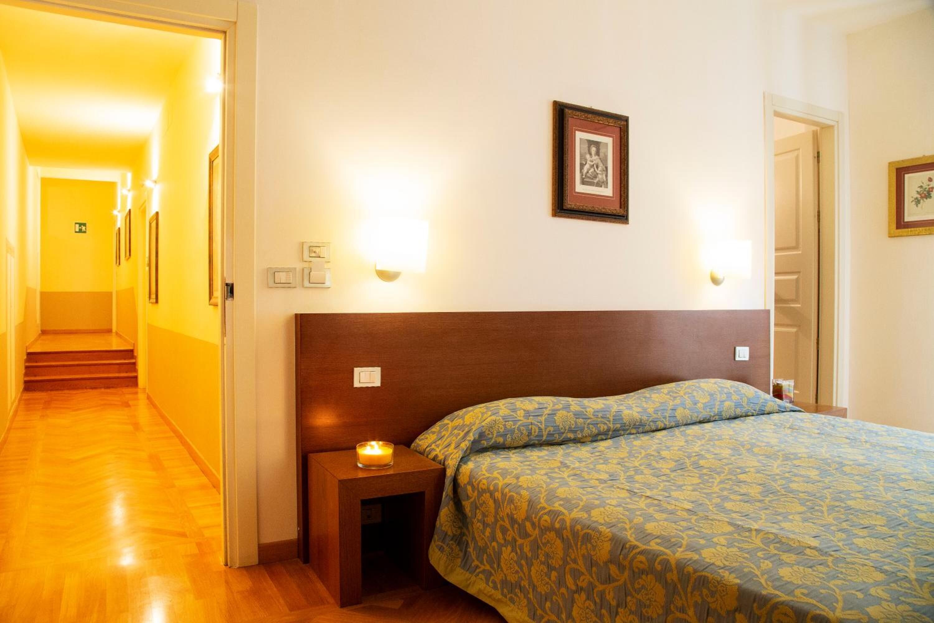 A hotel room with a wooden headboard, a bed with patterned bedding, a nightstand with a candle, and a framed picture on the wall.