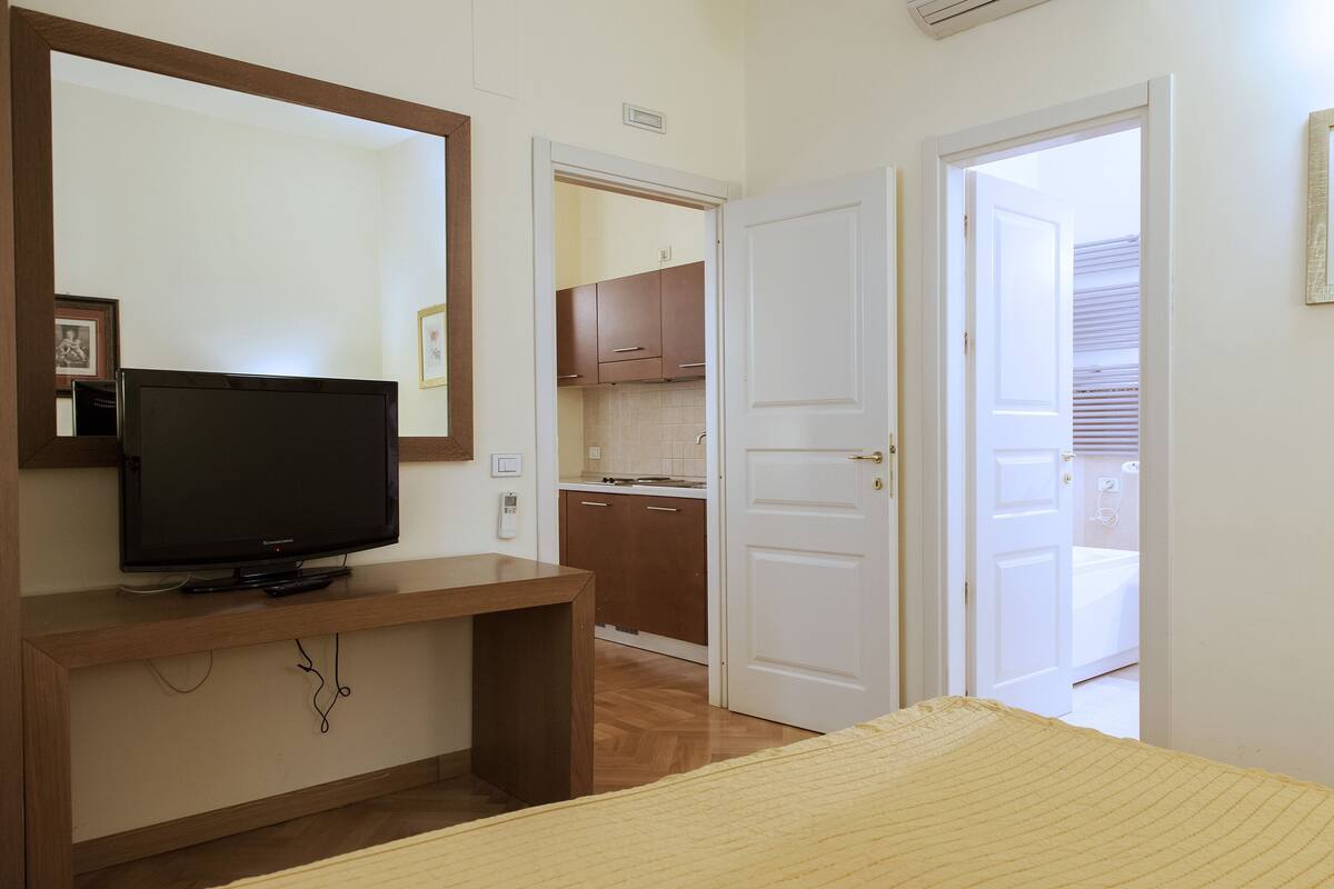 Une chambre avec un téléviseur posé sur un meuble en bois, un lit recouvert d’une couverture jaune, un miroir et une porte donnant sur une salle de bain.