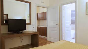 A bedroom with a TV on a wooden stand, a bed with a yellow blanket, a mirror, and a door leading to a bathroom.