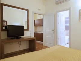 Quarto com televisão sobre suporte de madeira, cama com cobertor amarelo, espelho e porta que dá para um banheiro.