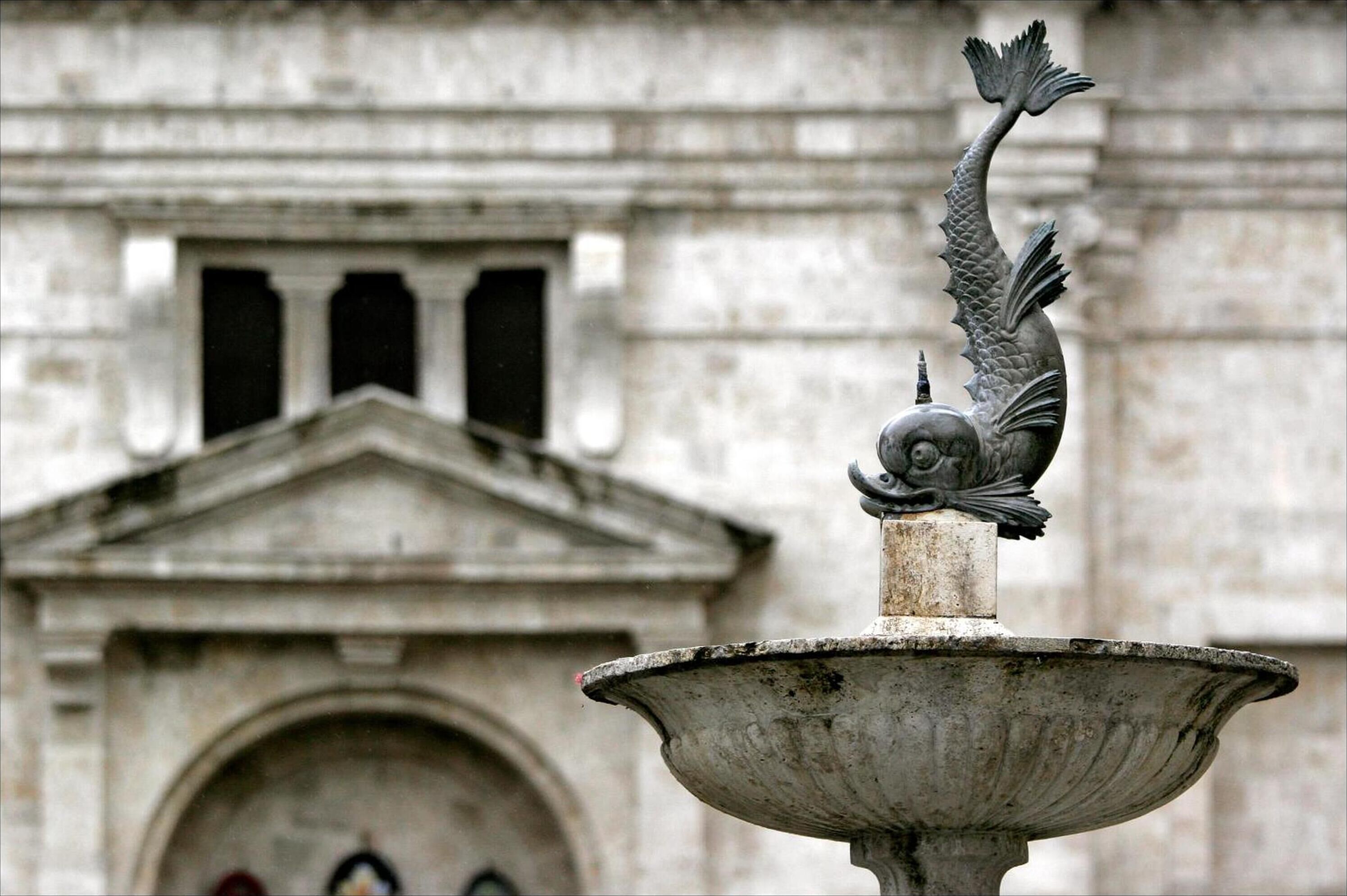 A stone fountain with a fish sculpture in front of a classical building facade.