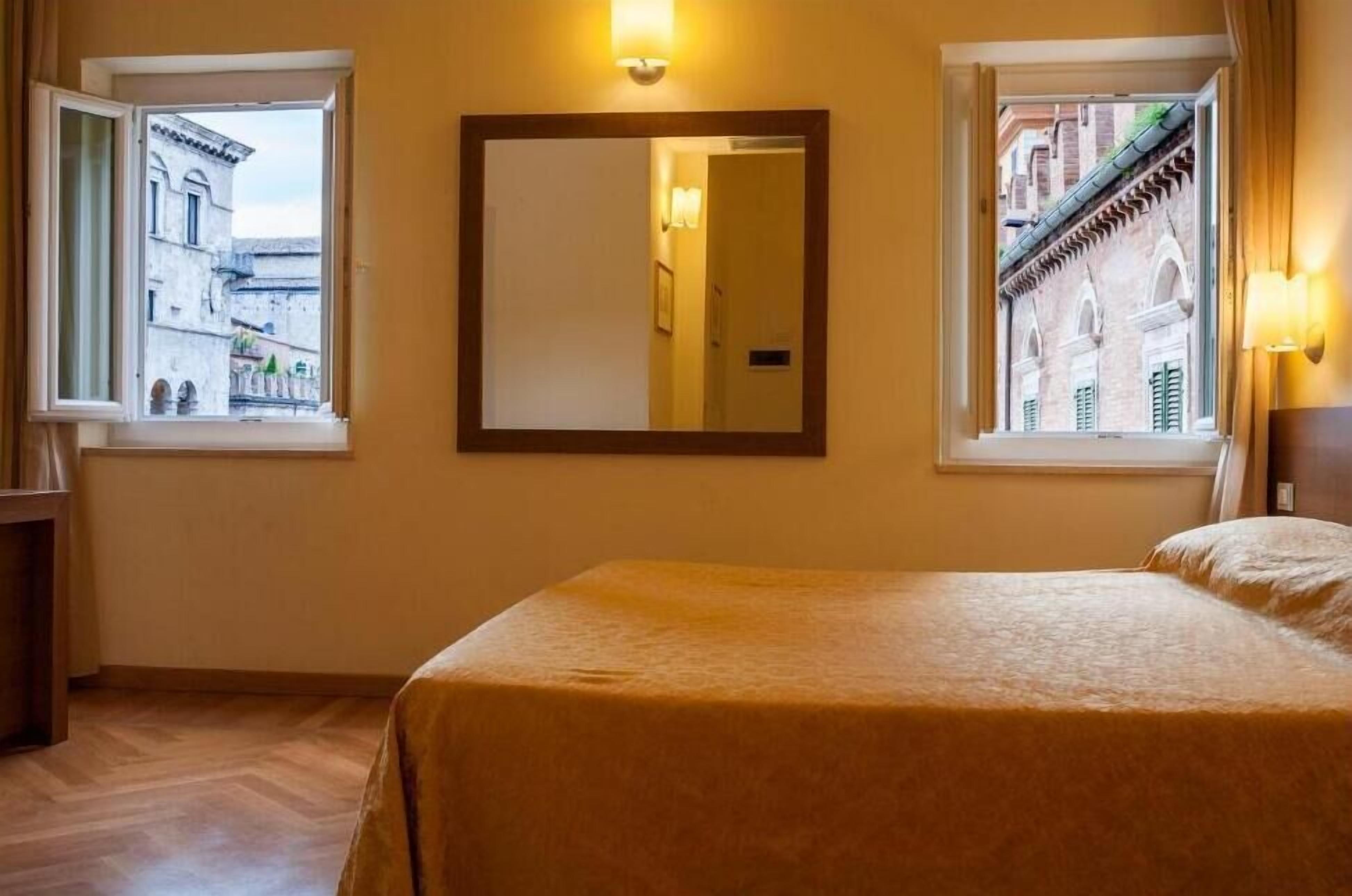 Ein Hotelzimmer mit einem Bett, zwei Fenstern mit Blick auf Gebäude, einem Spiegel und Wandleuchten.
