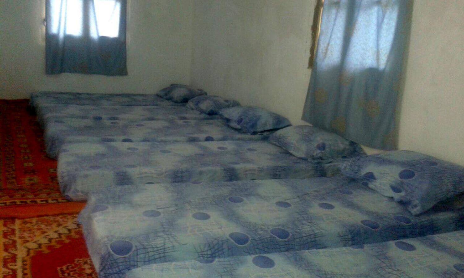 Una habitación con tres camas dispuestas en línea, una alfombra roja con estampado y una ventana con cortinas.