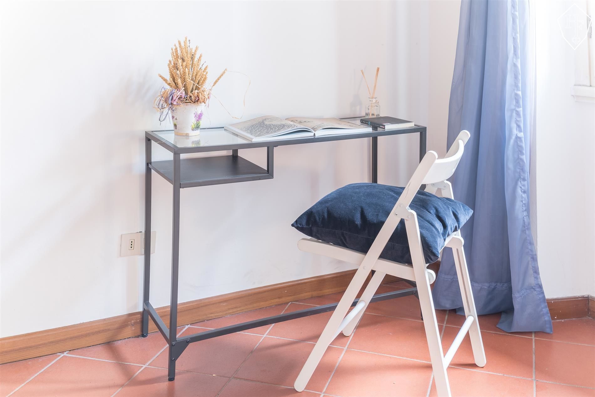 A small, modern workspace with a foldable chair, a glass-top desk, and a decorative vase with dried plants.