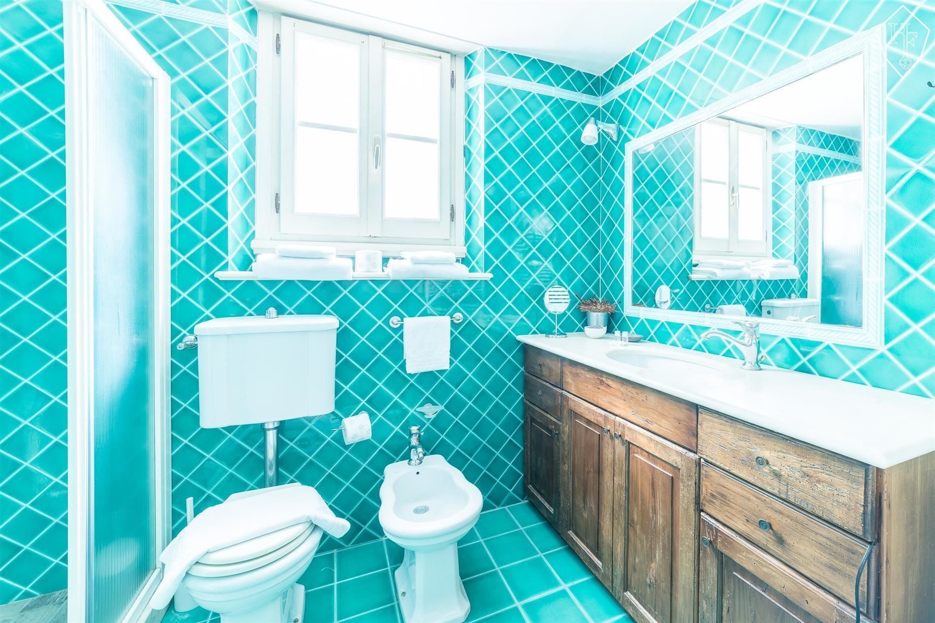 Een badkamer met toilet, bidet en wastafel, met turquoise tegels en houten kastjes.
