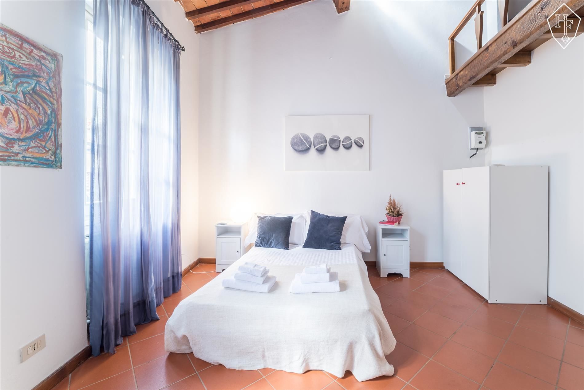 A bedroom with a bed, white curtains, a painting, a wall-mounted artwork, a white wardrobe, and a tiled floor.