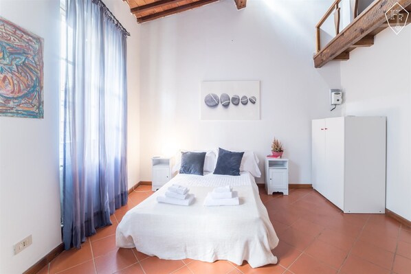 Un dormitorio con cama, cortinas blancas, un cuadro, una obra de arte en la pared, un armario blanco y suelo de baldosas.