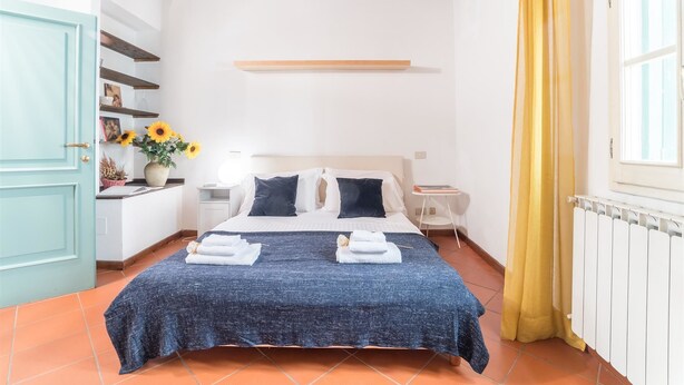Un dormitorio con cama, mesitas de noche, una ventana con cortinas y un estante en la pared.