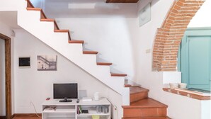 Una escalera con paredes blancas y escalones de madera que lleva a una habitación con una televisión, estanterías y una pared de ladrillo.