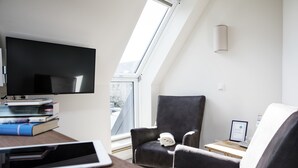 A modern living room with a flat-screen TV, a stack of books, a tablet, a grey armchair, and a wooden side table. - Hotel Jess am Meer (Büsum)