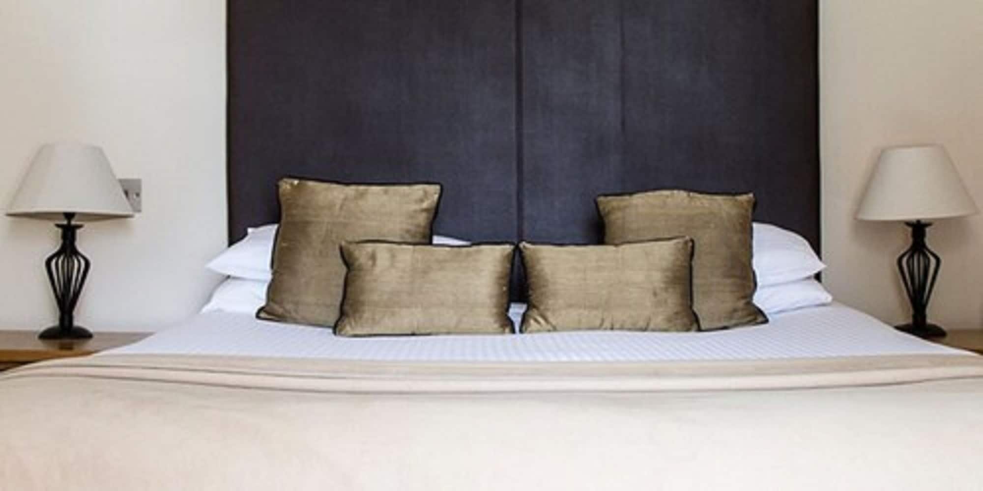 A bed with a dark headboard, four beige pillows, white linens, and two bedside lamps on either side.
