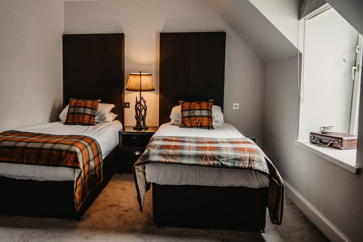 A hotel room with two beds, a nightstand with a lamp, a window with a suitcase, and a wall clock.