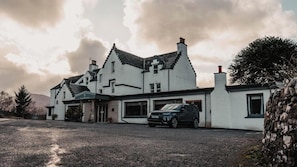 Un edificio blanco grande con varias chimeneas, un garaje con un coche y un camino de entrada de grava.