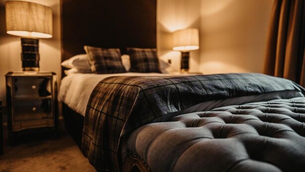 A neatly made bed with a plaid bedspread and a tufted headboard, flanked by two bedside tables with lamps. - Broadford Hotel (Isle of Skye)