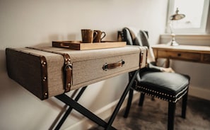Una maleta antigua reutilizada como mesita de noche con una bandeja y una taza encima, colocada junto a un escritorio y una silla en una habitación.