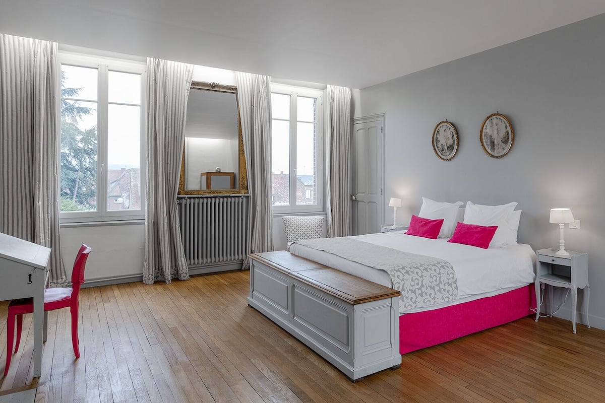 Quarto com cama, mesas de cabeceira, uma cadeira, espelho e janela com cortinas.