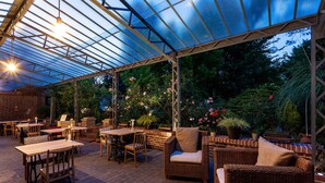 An outdoor patio with tables and wicker chairs, surrounded by greenery and a glass roof structure.