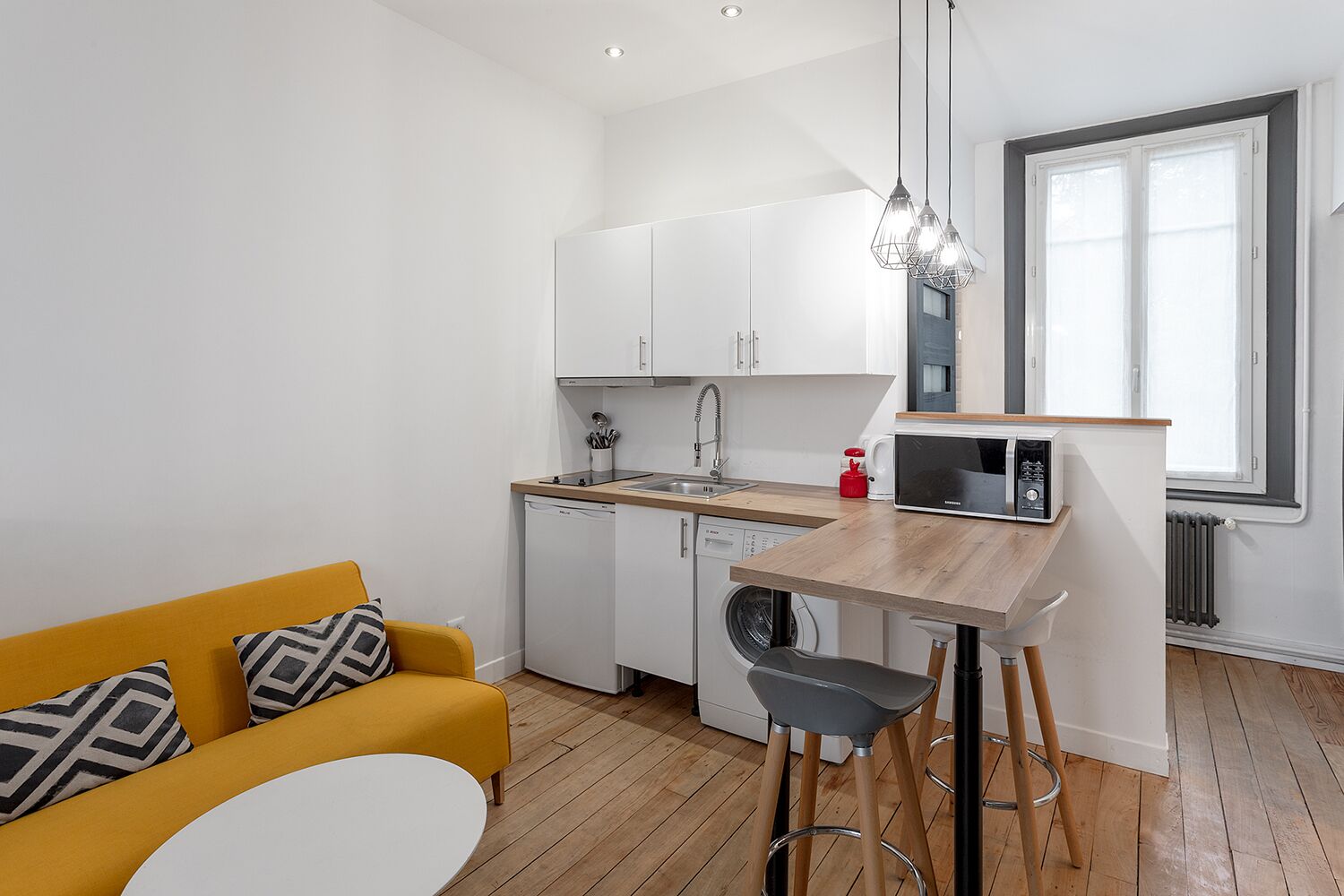 Una cocina compacta con armarios blancos, un microondas y un lavamanos. Hay un sofá amarillo con cojines estampados y una pequeña mesa redonda.