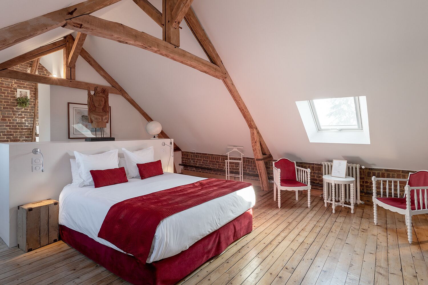 Un dormitorio con una cama grande, vigas de madera, pared de ladrillo, una ventana y dos sillas blancas.