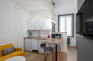 Cocina compacta con armarios blancos, un microondas y un lavamanos. Hay una silla amarilla y una pequeña mesa con taburetes.
