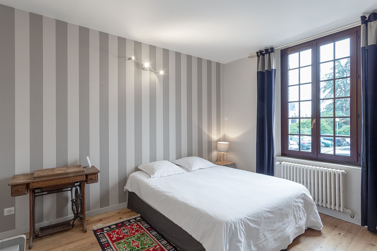 A bedroom with a bed, a nightstand, a lamp, a radiator, and a window with curtains.