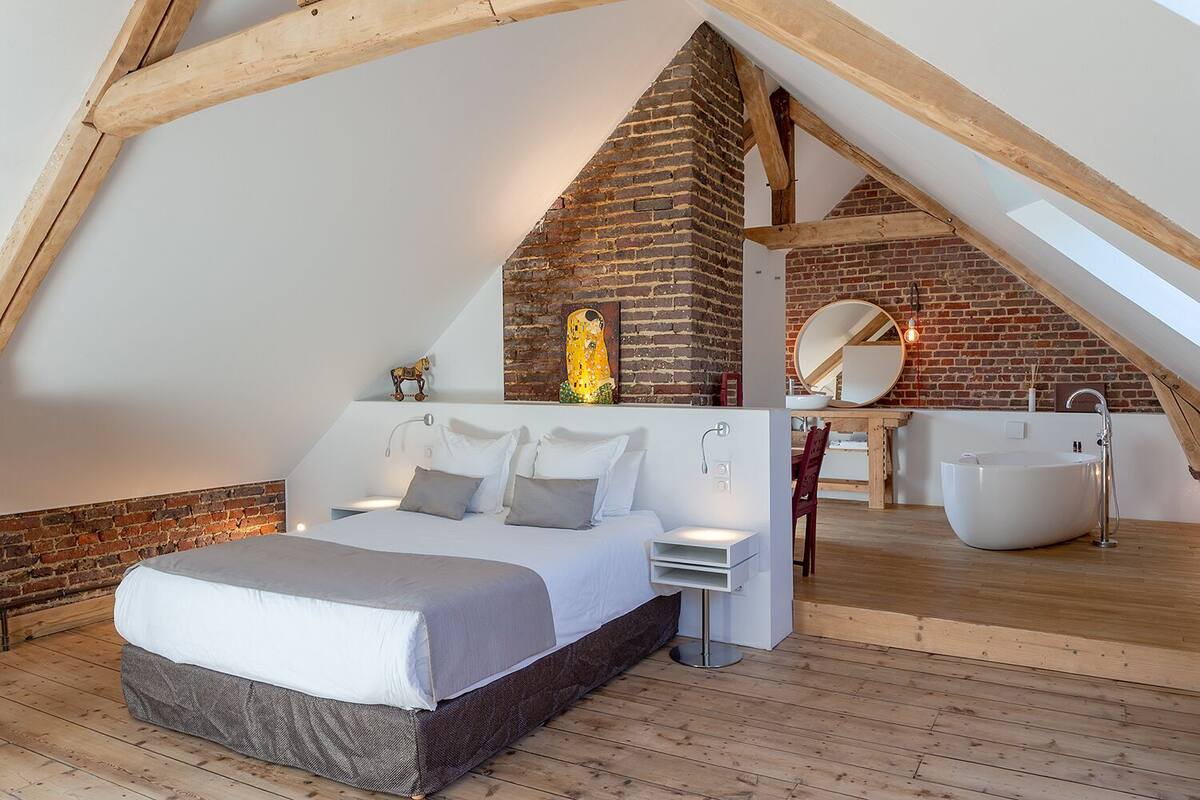 Quarto com teto inclinado, parede de tijolos, cama, mesas de cabeceira, espelho e banheira integrada.