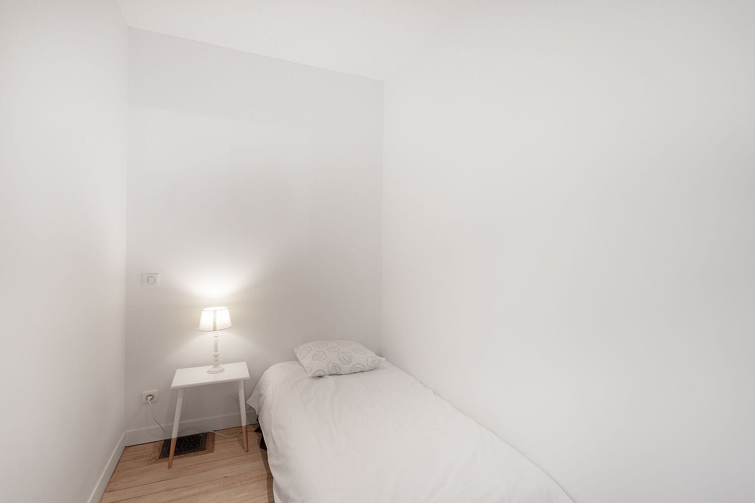 Una habitación pequeña con una cama individual, una mesita de noche y una lámpara.