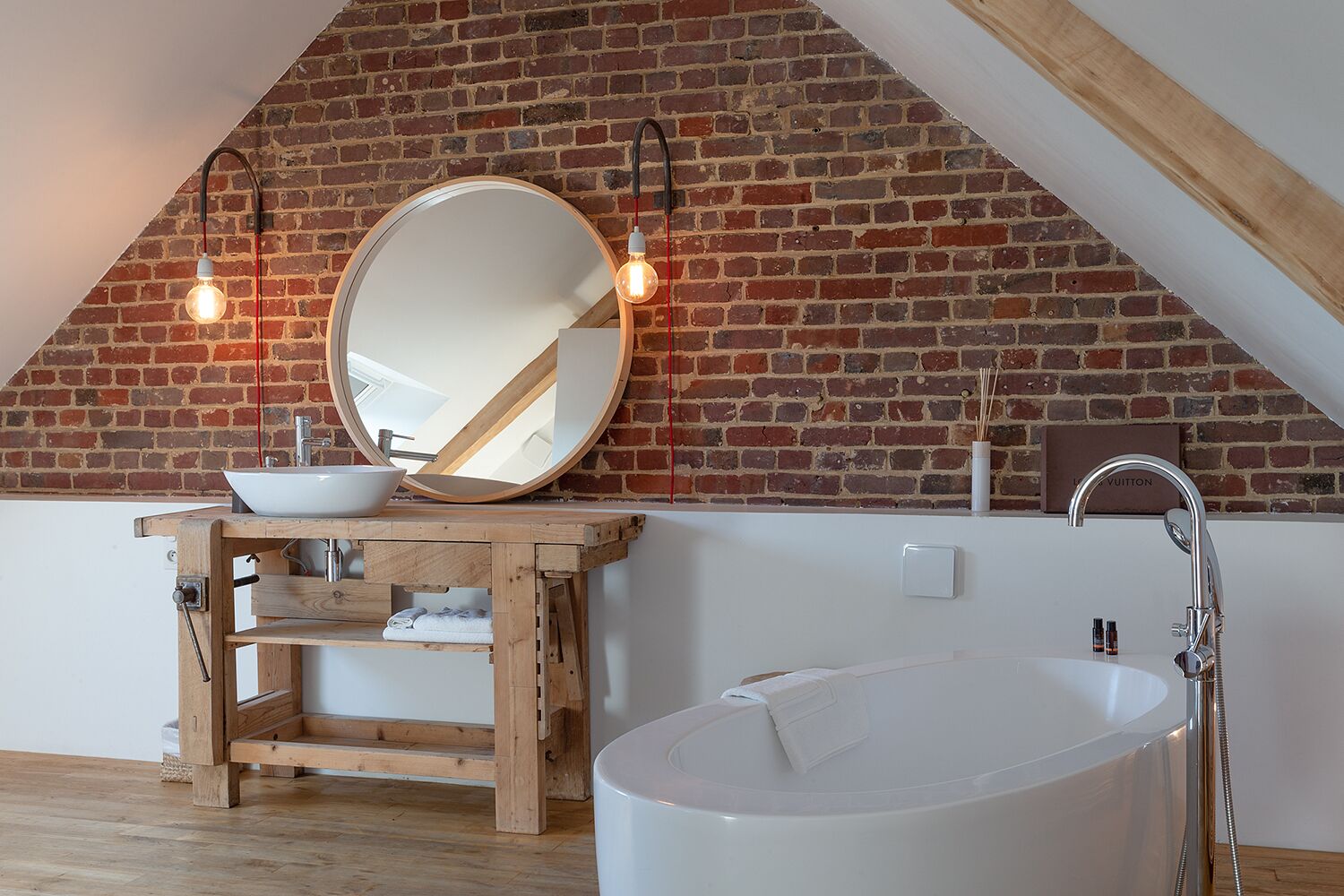 Une salle de bain avec une baignoire blanche, un meuble-lavabo en bois avec un vasque, un grand miroir et un mur en briques.