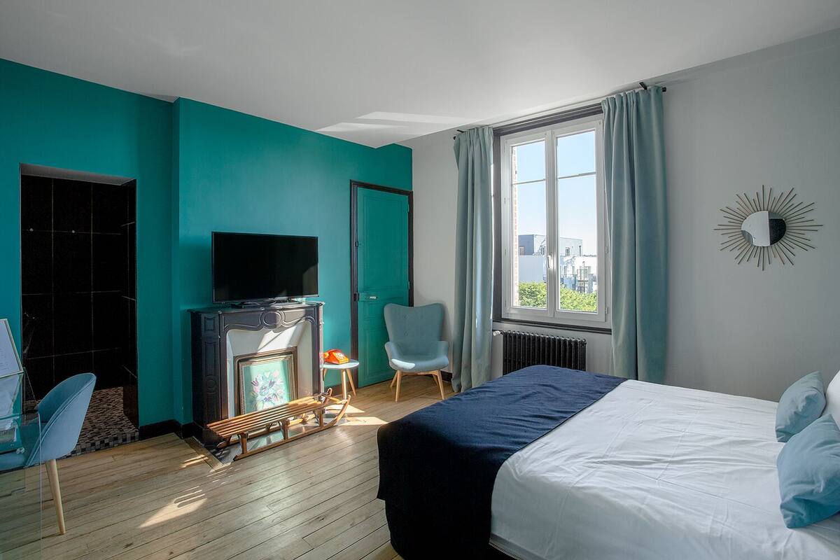 Quarto de hotel moderno com paredes verde-água, uma cama, uma televisão, lareira e vista para a cidade pela janela.