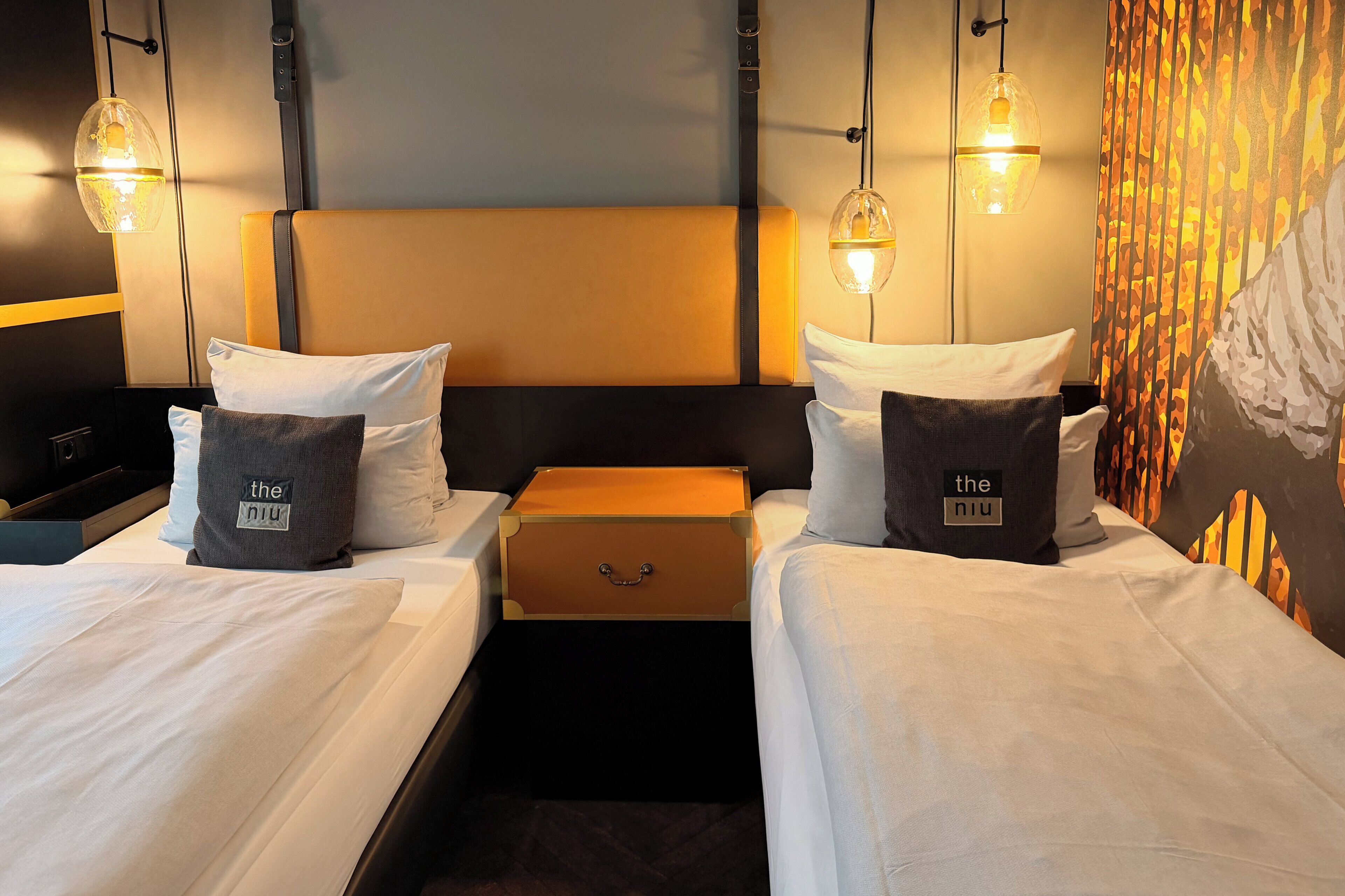 Quarto de hotel com duas camas, uma mesinha de cabeceira e uma mala.