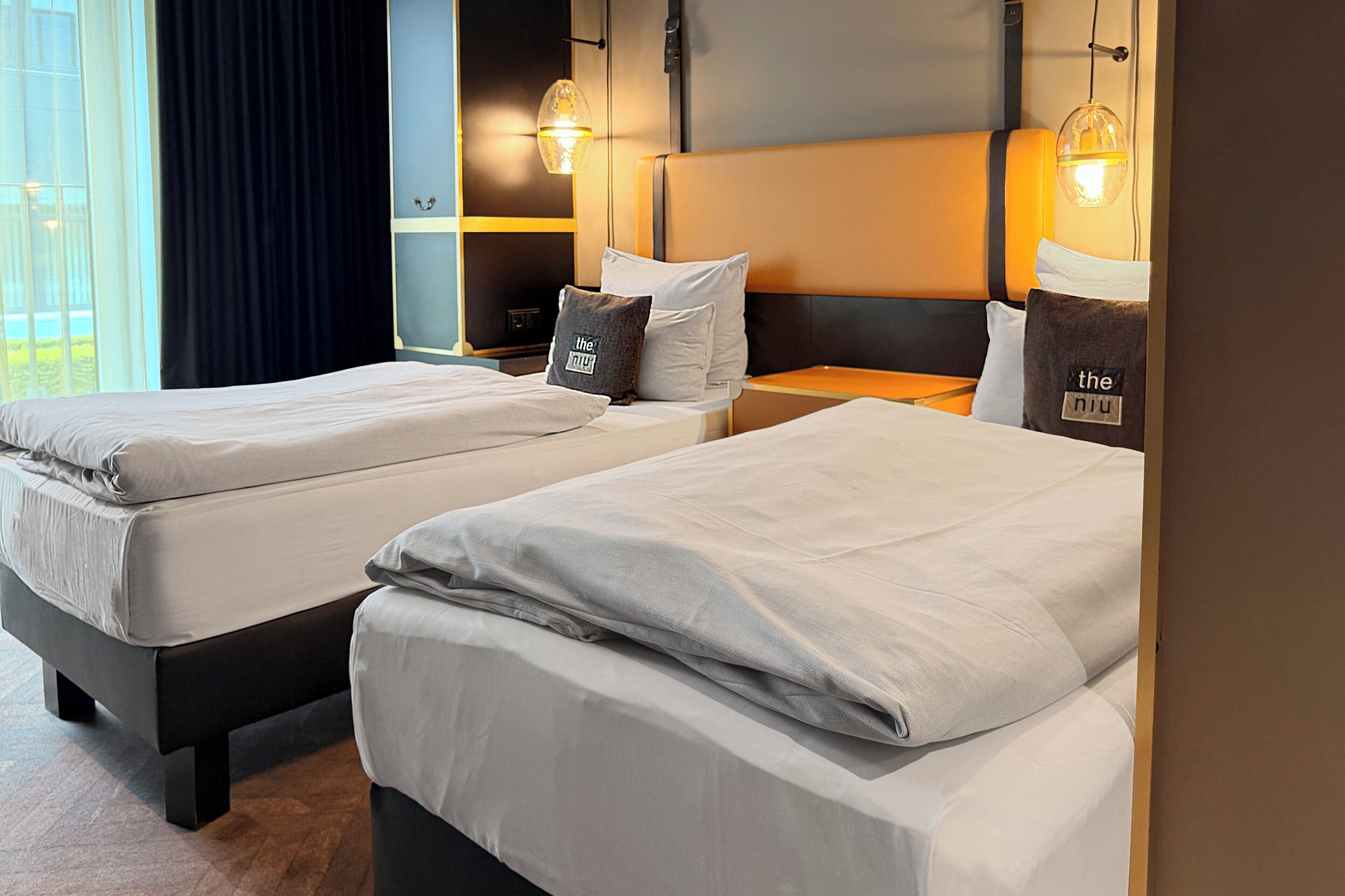 Habitación de hotel con dos camas, una mesita de noche, una lámpara y vistas al exterior.