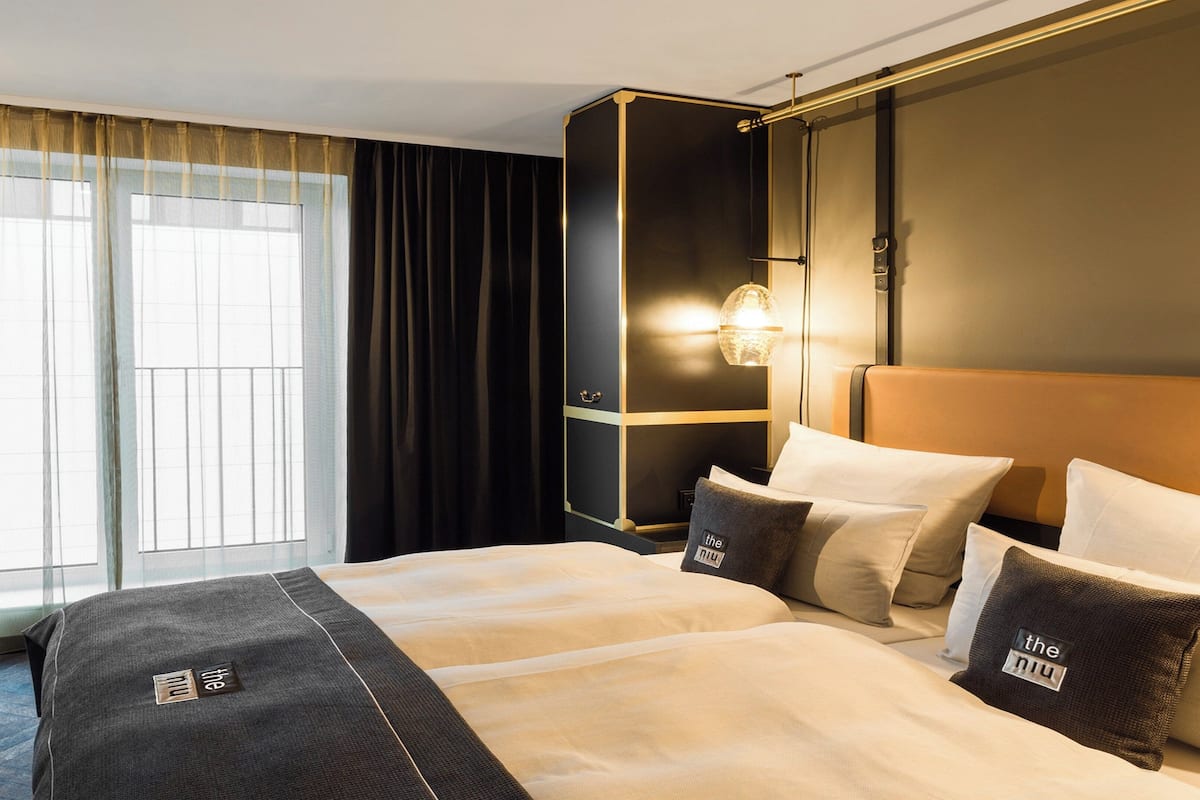 A hotel room with a large bed, a nightstand, a wall-mounted lamp, and a window with curtains.