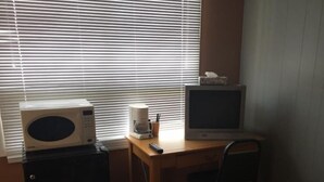 Um quarto pequeno com uma escrivaninha de madeira, micro-ondas, máquina de café, televisão e uma cadeira.